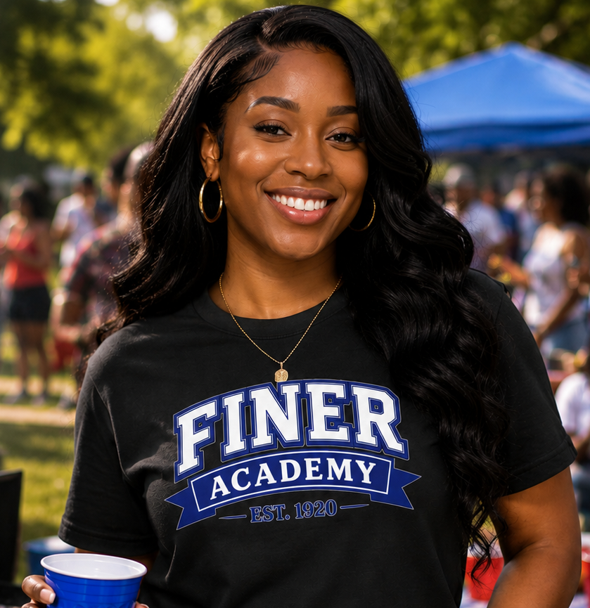 Woman wearing a black t-shirt with 'FINER ACADEMY' text, holding a blue cup, at an outdoor event.